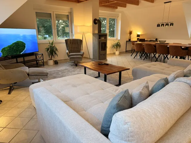 BookNoFees: Ferienwohnung in Stuhr – Wohnzimmer mit L-förmigem Sofa, Holzofen und Essbereich mit Holztisch