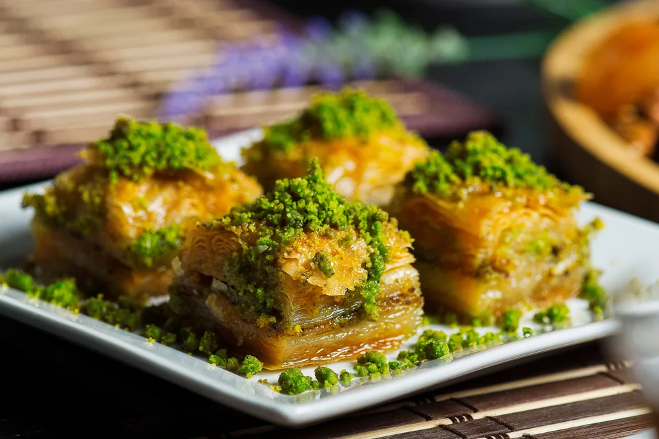 BookNoFees: Beispielfoto: Vier Stück geschichtetes Baklava, garniert mit zerkleinerten Pistazien, auf einem weißen Teller